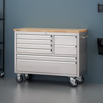 A stainless steel mobile tool workbench with a solid light wood top rests against a blue-gray wall in a workshop setting. The unit features all-closed drawers, four heavy-duty caster wheels, and a side pull handle, flanked by a black wire shelving unit on the left and wall-mounted cleaning tools on the right.