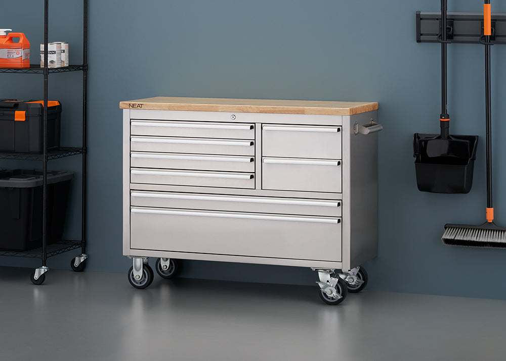A stainless steel mobile tool workbench with a solid light wood top rests against a blue-gray wall in a workshop setting. The unit features all-closed drawers, four heavy-duty caster wheels, and a side pull handle, flanked by a black wire shelving unit on the left and wall-mounted cleaning tools on the right.