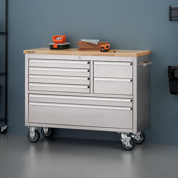 A stainless steel mobile tool workbench with a light wood top is shown in a workshop against a blue-gray wall, with all drawers securely closed. Small hardware bins, wood blocks, and a tape measure rest on top, flanked by a black wire shelving unit on the left and wall-mounted cleaning tools on the right.