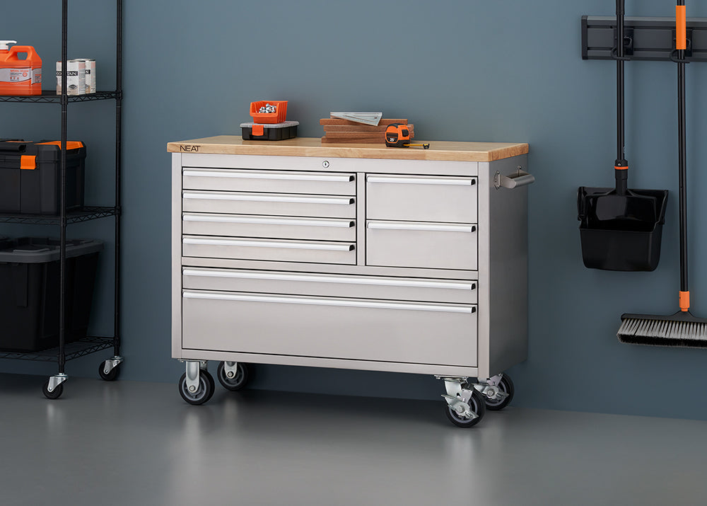 A stainless steel mobile tool workbench with a light wood top is shown in a workshop against a blue-gray wall, with all drawers securely closed. Small hardware bins, wood blocks, and a tape measure rest on top, flanked by a black wire shelving unit on the left and wall-mounted cleaning tools on the right.