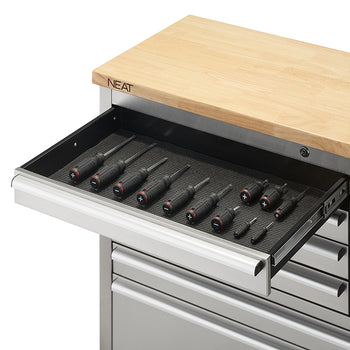 A close-up view focuses on the top-left shallow drawer of a stainless steel tool workbench, extended on metal slides to show black-handled screwdrivers organized neatly on a textured drawer liner. The light wood top above displays the "NEAT" logo branded on its corner edge.