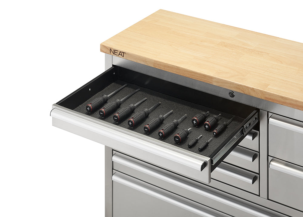 A close-up view focuses on the top-left shallow drawer of a stainless steel tool workbench, extended on metal slides to show black-handled screwdrivers organized neatly on a textured drawer liner. The light wood top above displays the "NEAT" logo branded on its corner edge.