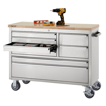 A stainless steel mobile tool workbench with a light wood top rests on swivel casters against a plain white background. One of the upper drawers on the left side is pulled open to show small organizer boxes stored inside, while a cordless drill, level, and small tool set rest on top of the wooden surface.