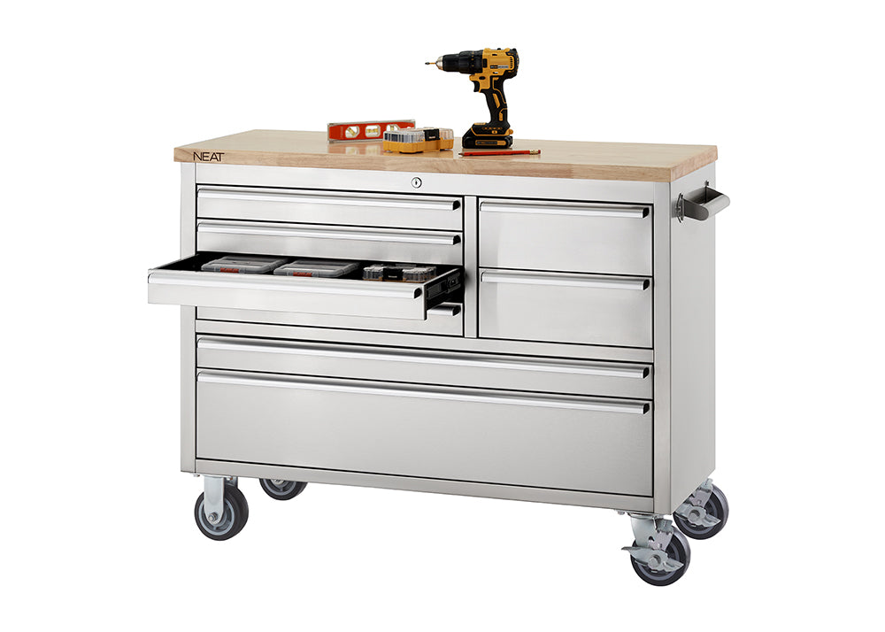 A stainless steel mobile tool workbench with a light wood top rests on swivel casters against a plain white background. One of the upper drawers on the left side is pulled open to show small organizer boxes stored inside, while a cordless drill, level, and small tool set rest on top of the wooden surface.