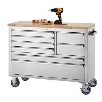 A stainless steel mobile tool workbench with a light wood top rests on swivel casters against a plain white background. The unit features multiple closed drawers of varying sizes, with the "NEAT" logo visible on the corner of the wooden top, and a cordless drill, level, and small tool set organized on the surface.