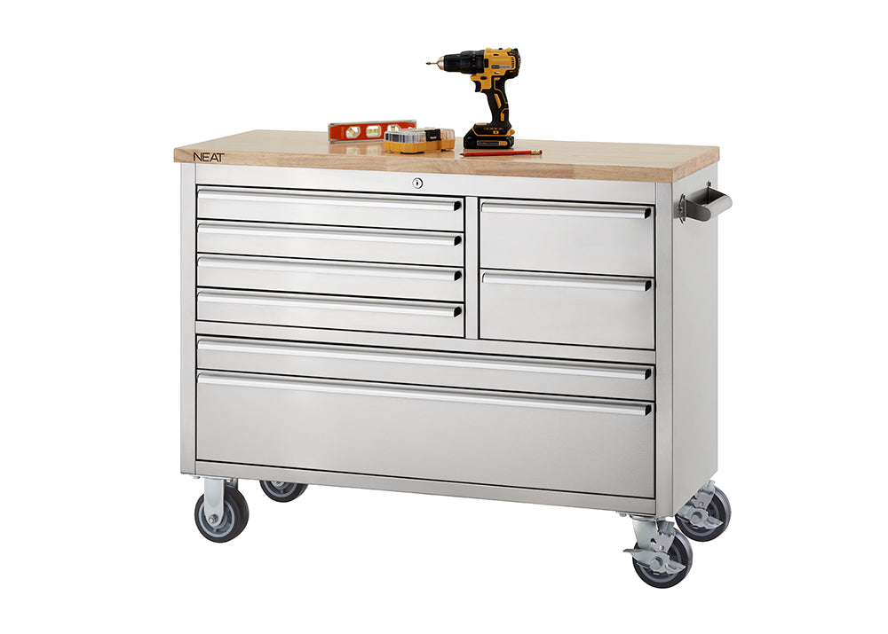 A stainless steel mobile tool workbench with a light wood top rests on swivel casters against a plain white background. The unit features multiple closed drawers of varying sizes, with the "NEAT" logo visible on the corner of the wooden top, and a cordless drill, level, and small tool set organized on the surface.
