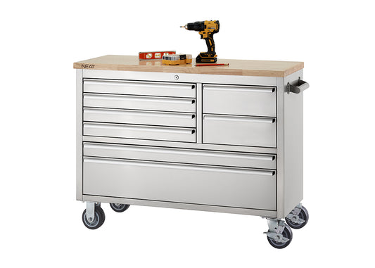 A stainless steel mobile tool workbench with a light wood top rests on swivel casters against a plain white background. The unit features multiple closed drawers of varying sizes, with the "NEAT" logo visible on the corner of the wooden top, and a cordless drill, level, and small tool set organized on the surface.