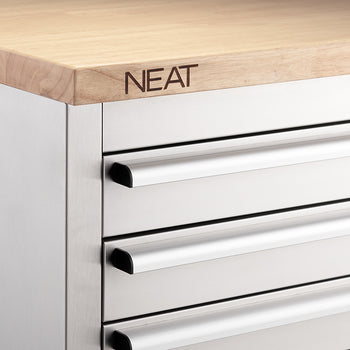 A close-up view focuses on the upper-left corner of a stainless steel tool workbench, highlighting the smooth metal drawer handles and the "NEAT" brand logo stamped into the edge of the light wood countertop.