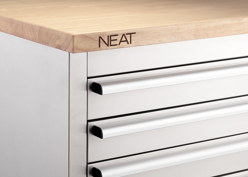 A close-up view focuses on the upper-left corner of a stainless steel tool workbench, highlighting the smooth metal drawer handles and the "NEAT" brand logo stamped into the edge of the light wood countertop.
