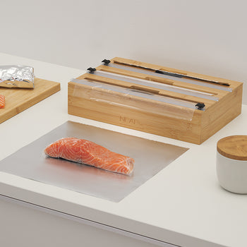 A bamboo wrap dispenser sits on a white countertop next to salmon fillets being prepared with foil and plastic wrap. The image illustrates the dispenser in use during meal prep, highlighting its ability to provide clean, precise cuts for food preservation.