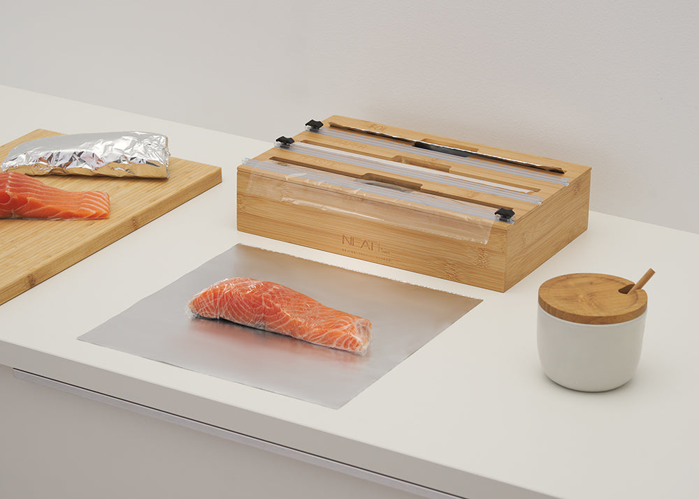 A bamboo wrap dispenser sits on a white countertop next to salmon fillets being prepared with foil and plastic wrap. The image illustrates the dispenser in use during meal prep, highlighting its ability to provide clean, precise cuts for food preservation.