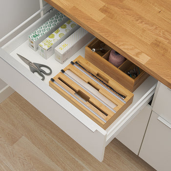 An open white kitchen drawer displays a bamboo wrap dispenser along with scissors, boxes of bags, and a wooden organizer for twine and clips. The setup demonstrates how the low-profile bamboo dispenser fits neatly inside a standard drawer for organized, out-of-sight storage.