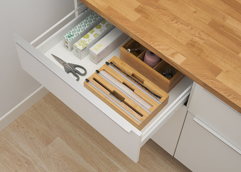 An open white kitchen drawer displays a bamboo wrap dispenser along with scissors, boxes of bags, and a wooden organizer for twine and clips. The setup demonstrates how the low-profile bamboo dispenser fits neatly inside a standard drawer for organized, out-of-sight storage.