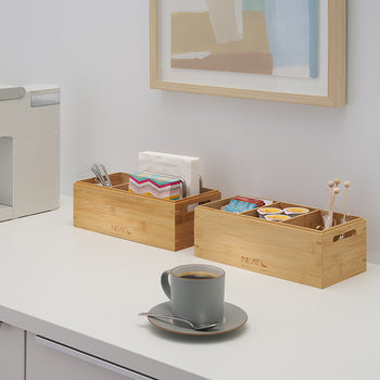 A gray coffee mug with a spoon rests on a white counter in front of a modern beverage station. To the side, a white single-serve coffee machine stands next to two bamboo organizers holding napkins, utensils, coffee pods, and stirrers.