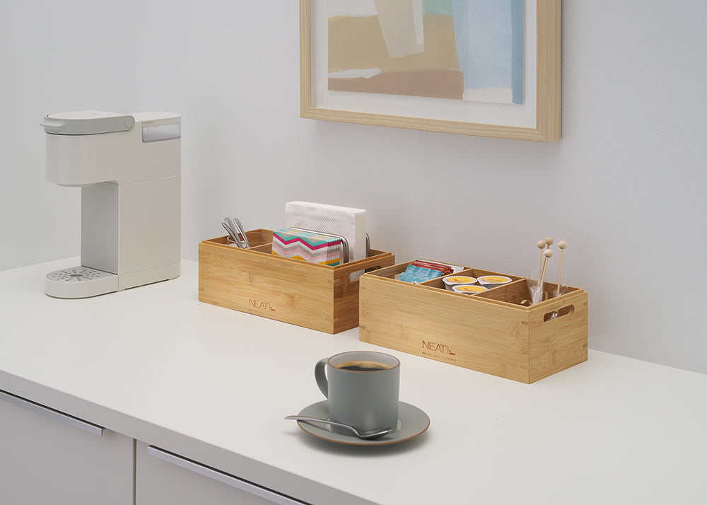 A gray coffee mug with a spoon rests on a white counter in front of a modern beverage station. To the side, a white single-serve coffee machine stands next to two bamboo organizers holding napkins, utensils, coffee pods, and stirrers.