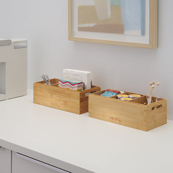A white single-serve coffee maker sits on a clean counter next to two bamboo organizers filled with napkins, stirrers, and coffee pods. Above them is a simple framed piece of art, completing the neat and organized beverage station.