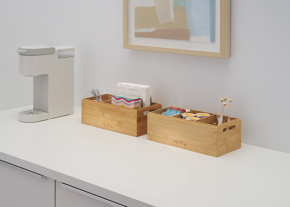 A white single-serve coffee maker sits on a clean counter next to two bamboo organizers filled with napkins, stirrers, and coffee pods. Above them is a simple framed piece of art, completing the neat and organized beverage station.