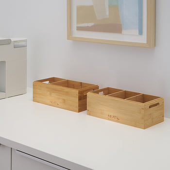 A white coffee pod maker stands on a white kitchen counter next to two empty bamboo boxes with multiple compartments and side handles. The bins are engraved with the word "NEAT" and positioned below a light wood framed piece of artwork against a light gray wall.