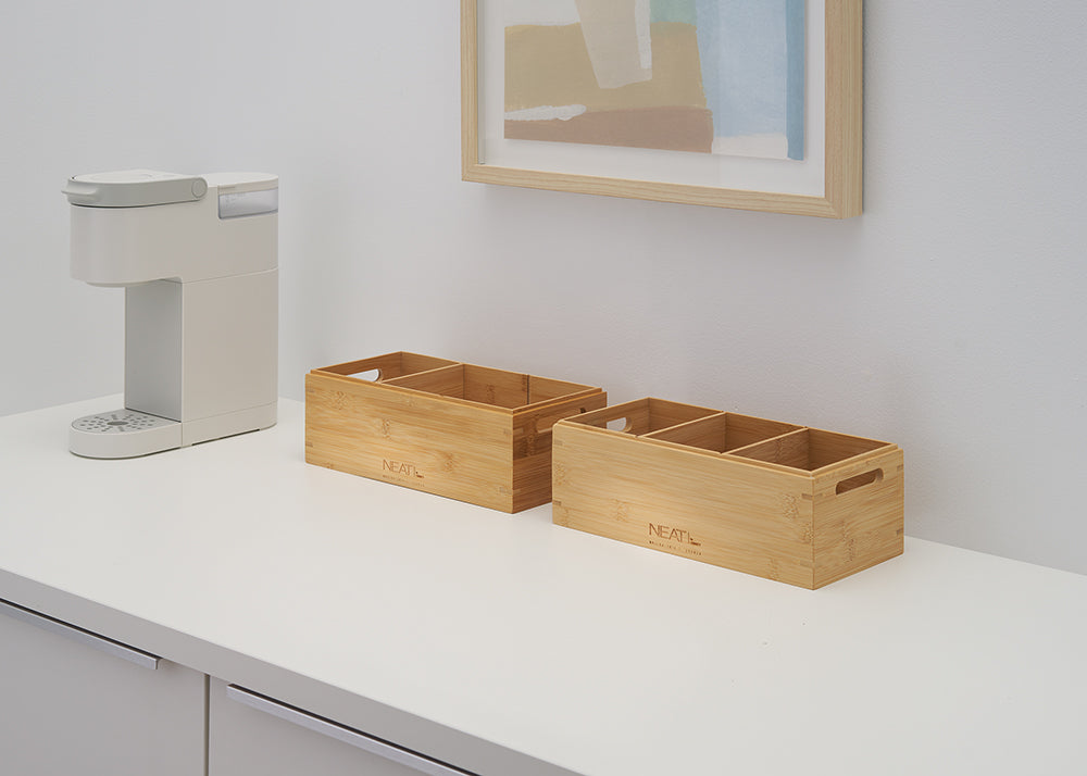 A white coffee pod maker stands on a white kitchen counter next to two empty bamboo boxes with multiple compartments and side handles. The bins are engraved with the word "NEAT" and positioned below a light wood framed piece of artwork against a light gray wall.