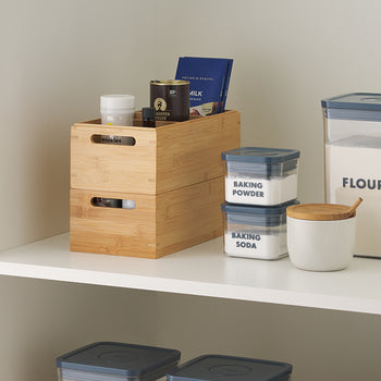 A stacked pair of bamboo bins holds assorted spice jars and boxes, next to clearly labeled clear plastic containers for flour, baking powder, and baking soda. A white ceramic jar with a bamboo lid and small wooden spoon rests nearby on the clean white pantry shelf.