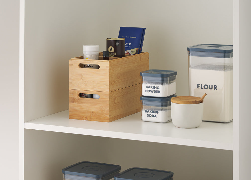A stacked pair of bamboo bins holds assorted spice jars and boxes, next to clearly labeled clear plastic containers for flour, baking powder, and baking soda. A white ceramic jar with a bamboo lid and small wooden spoon rests nearby on the clean white pantry shelf.