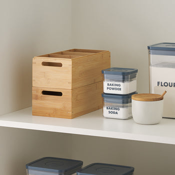 A white pantry shelf displays a set of stacked bamboo storage boxes with cutout handles and four compartments. Next to them are labeled airtight containers for flour, baking powder, and baking soda, along with a white ceramic jar and wooden spoon.