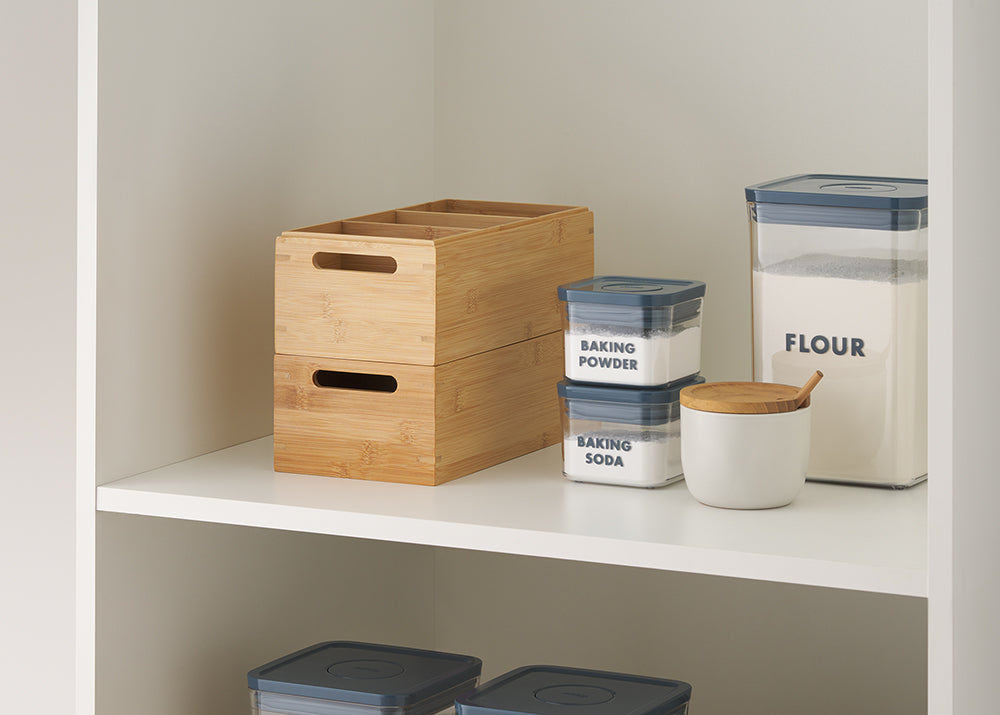 A white pantry shelf displays a set of stacked bamboo storage boxes with cutout handles and four compartments. Next to them are labeled airtight containers for flour, baking powder, and baking soda, along with a white ceramic jar and wooden spoon.