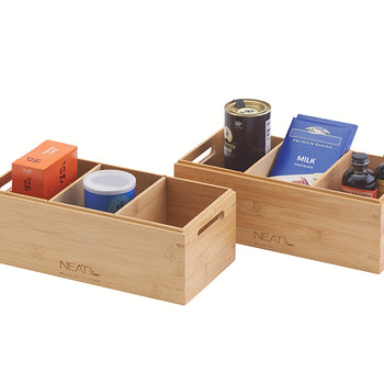A front three-quarter view displays two natural bamboo storage bins, one wider with four internal compartments and a smaller one with two, positioned against a plain white background. The bins are filled with assorted pantry items including spice tins, cocoa powder, chocolate bars, and glass jars, and feature integrated handles and "NEAT" branding.