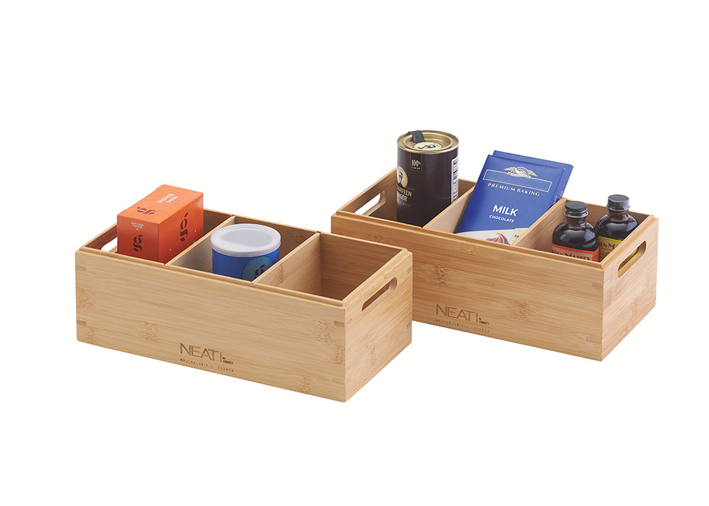 A front three-quarter view displays two natural bamboo storage bins, one wider with four internal compartments and a smaller one with two, positioned against a plain white background. The bins are filled with assorted pantry items including spice tins, cocoa powder, chocolate bars, and glass jars, and feature integrated handles and "NEAT" branding.