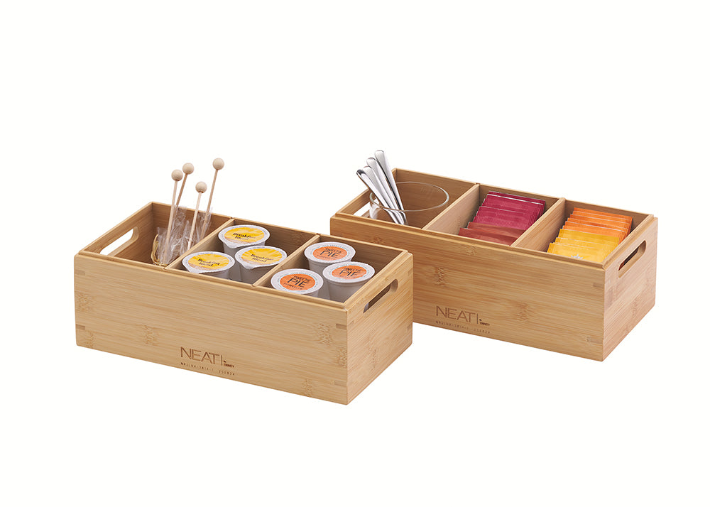 A front three-quarter view shows two natural bamboo storage organizers with four and three compartments respectively, positioned against a plain white background. Inside, they are neatly packed with various coffee and tea supplies, including K-Cups, stirrers, spice tins, and tea bags, and feature integrated handles for easy transport.
