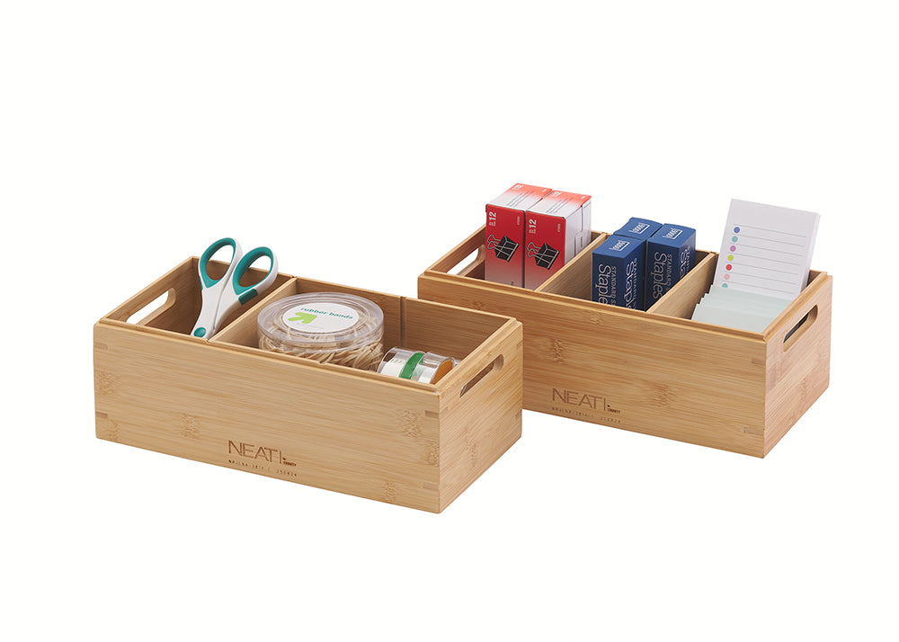 A natural wood multi-compartment desktop organizer is filled with various office and craft supplies, including scissors, a container of rubber bands, paper clips, staples, and note pads, against a plain white background. The organizer has integrated side handles for easy carrying.