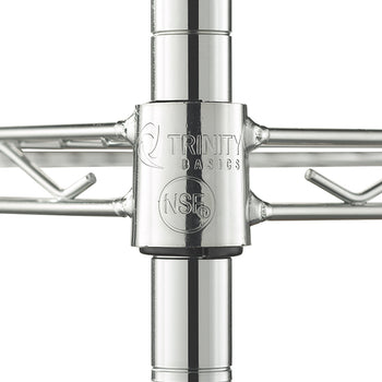 A close-up photograph focuses on a chrome metal leg joint from a wire shelf. The cylindrical connector is stamped with the "Trinity Basics" logo and a circular "NSF" mark, and it connects vertical posts with horizontal wire shelving that extends to the left and right.