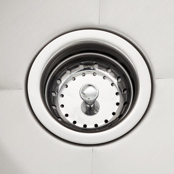 A close-up photograph shows the stainless steel drain basket of a kitchen sink, set against the smooth, shiny basin. The round basket has a pattern of holes for drainage and features a small, central metal knob, likely for removal or sealing the drain.