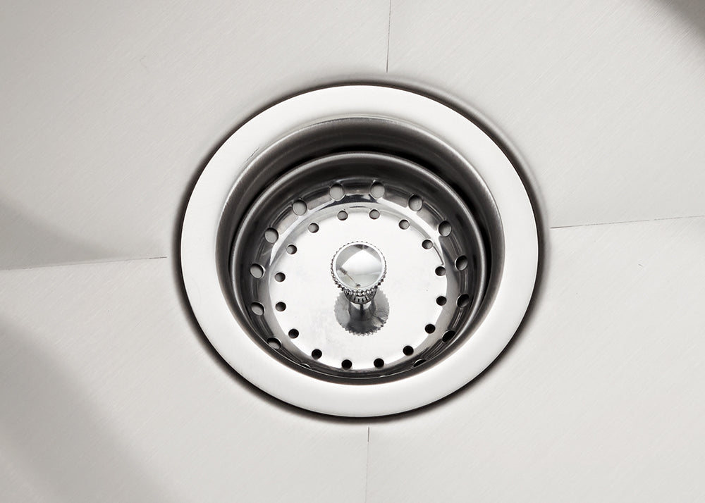 A top-down, close-up shot shows a polished stainless steel kitchen sink drain with a removable basket strainer in the center. The surrounding sink surface features a brushed metal finish with subtle diagonal drainage lines leading toward the circular opening.