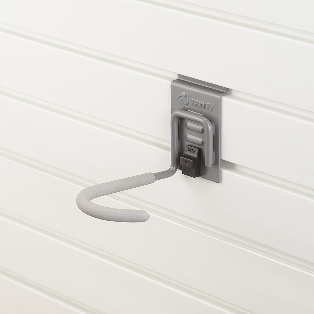 A gray metal utility hook with a foam-padded end is securely mounted on a white slatwall panel. The hook, branded "TRINITY," features a locking mechanism labeled "OPEN" for secure attachment and removal.