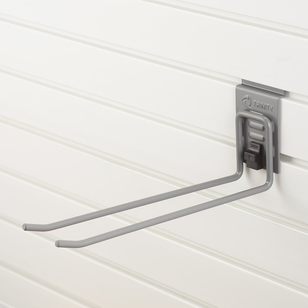 A gray metal double-prong hook is mounted on a white slatwall panel. The hook is branded "TRINITY" and features a locking mechanism labeled "OPEN" for secure attachment.