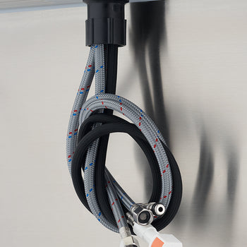 A close-up view from below shows a set of braided, flexible water supply hoses and a black pull-out hose hanging beneath a stainless steel surface, likely for a faucet installation. The hoses feature metal connectors and a distinctive white and orange quick-connect fitting.
