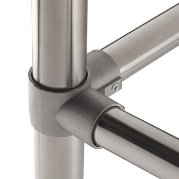 A close-up photograph shows a brushed metal joint connecting three round, stainless steel tubes at a right angle, forming a corner. The joint is secured with a small visible hex head screw, highlighting a modular pipe-fitting system.