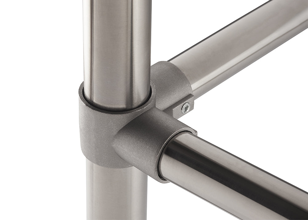 A close-up photograph shows a brushed metal joint connecting three round, stainless steel tubes at a right angle, forming a corner. The joint is secured with a small visible hex head screw, highlighting a modular pipe-fitting system.