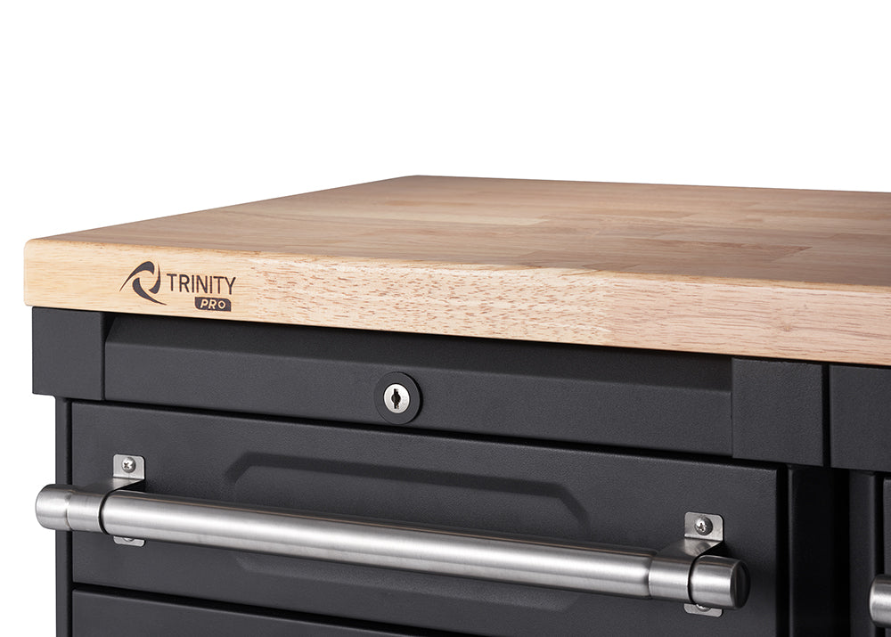 A close-up shot shows the top right corner of a black metal workbench with a light-colored wooden countertop. A "TRINITY PRO" logo is visible on the wood, and a keyhole is centered below it, above a long, silver drawer handle.