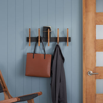 A black horizontal wall-mounted rack with five light brown wooden pegs hangs on a light blue paneled wall. A brown tote bag and a gray jacket are hanging from two of the pegs.