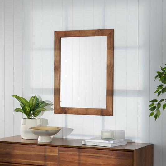 A rectangular walnut-framed mirror hangs on a white wall above a matching wooden dresser.  A green plant in a white pot sits to the left of the mirror, and a small decorative bowl and candle sit on the dresser.