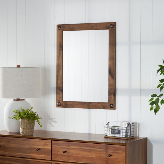 A rustic farmhouse style bathroom features a walnut-framed mirror with decorative clavos mounted above a matching dresser. The space is bright and airy, illuminated by a lamp on the dresser and complemented by the white shiplap wall.