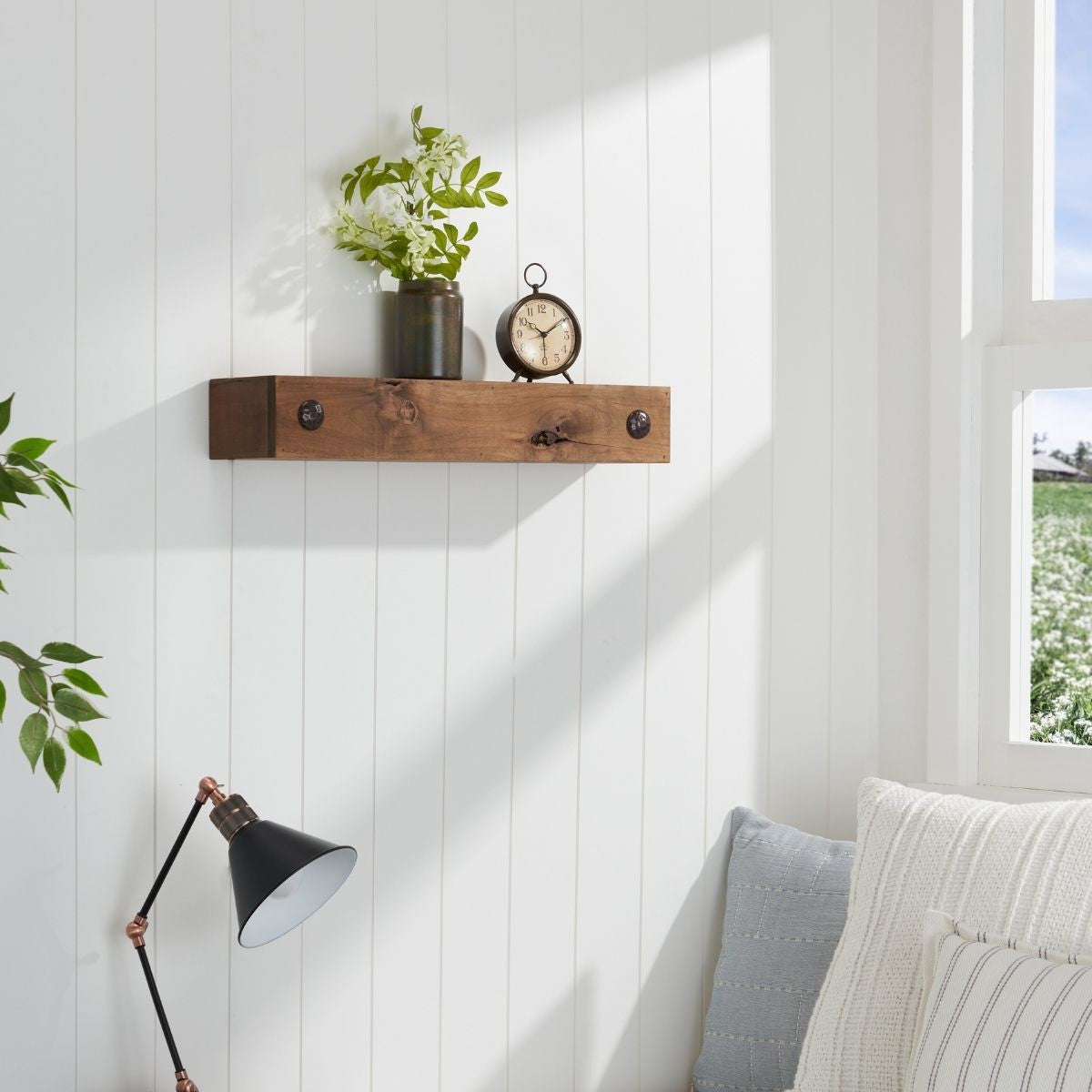 A walnut floating shelf with clavos is mounted on a white shiplap wall, holding a small clock and a vase of greenery.  A black and copper desk lamp is visible in the foreground, with a portion of a white sofa and pillows.