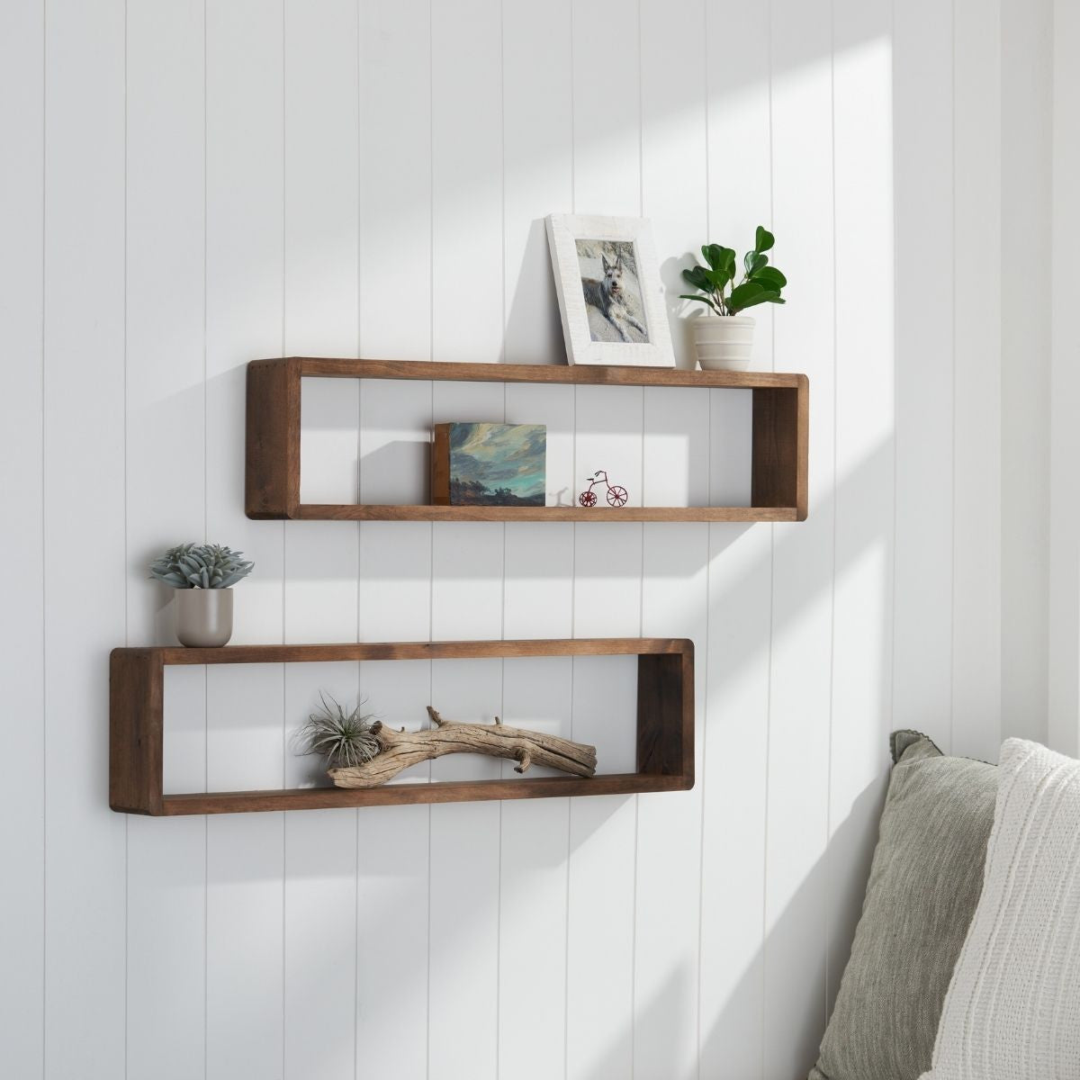Two rectangular wooden floating shelves are mounted on a white paneled wall, each displaying decorative items. The top shelf holds a framed picture, a small potted plant, a postcard, and a miniature bicycle, while the bottom shelf features a succulent in a gray pot and a piece of driftwood.
