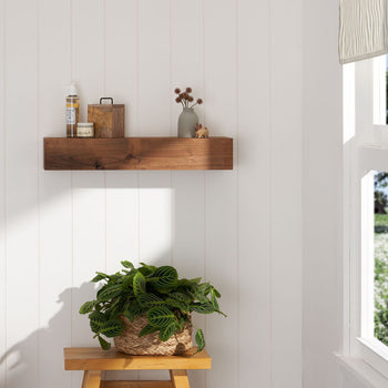 A wooden floating shelf is mounted on a white paneled wall, holding a few small decorative items. A potted plant sits on a wooden stool below the shelf, with a window to the right.