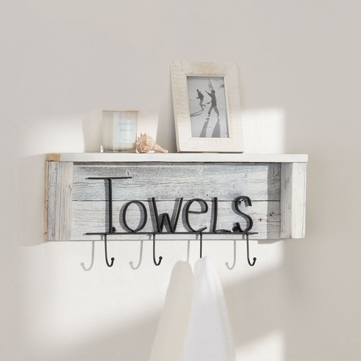 A rustic white wooden shelf with the word "Towels" in decorative black metal lettering hangs on a light beige wall. Below the shelf are five black metal hooks, one of which holds a folded white towel.