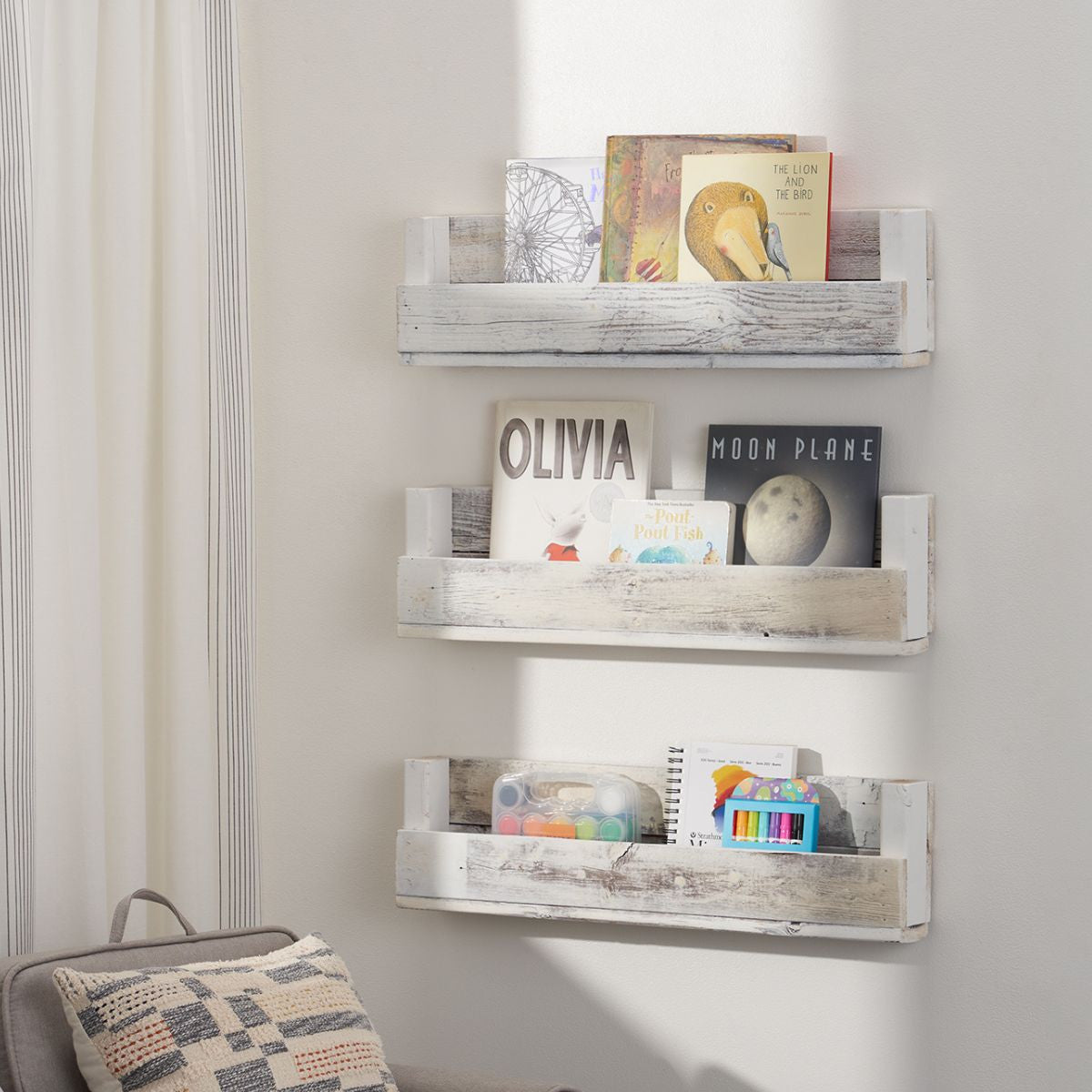 Three-tiered wall-mounted shelves made of rustic, reclaimed wood are styled as open bookshelves holding children's books and art supplies. The shelves are staggered vertically against a white wall in a room with a gray armchair and striped curtains.