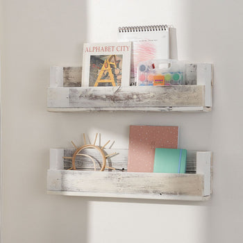 Two rustic white wooden wall shelves are mounted on a white wall, holding books, notebooks, and decorative items. The top shelf contains a book titled 