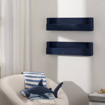 Two dark blue floating shelves are mounted on a white wall in a child's room, holding books facing forward. A white sherpa chair with a blue striped blanket and a shark plush toy sits in front of the shelves, with a window to the left showing a green field.
